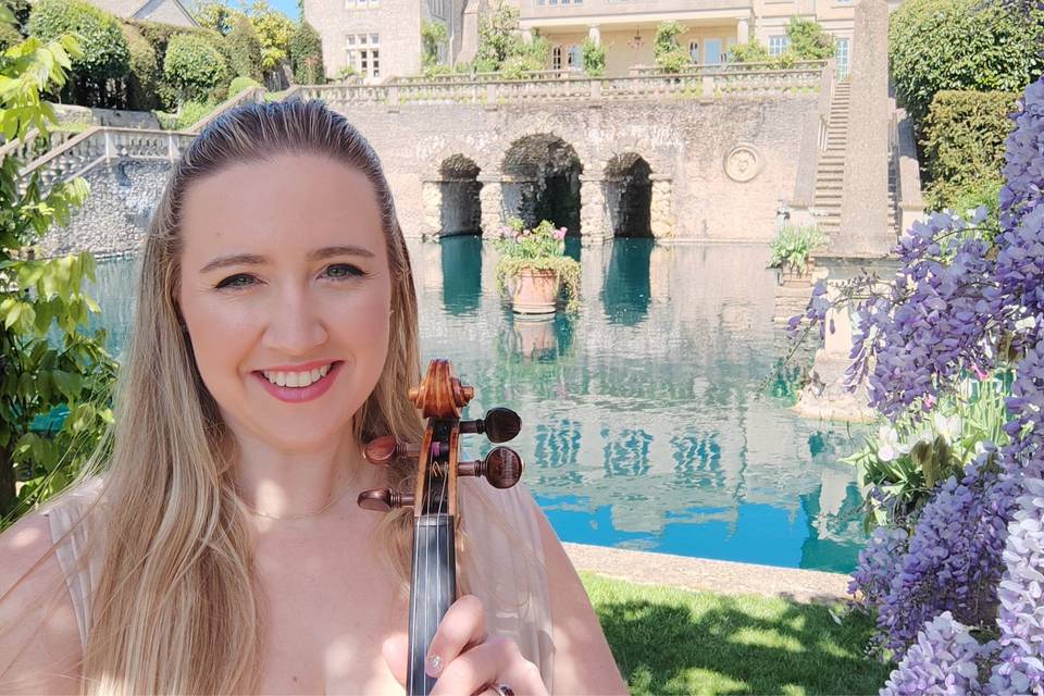 Smiling woman holding a violin by a pond with stone arches and flowering plants in the background.