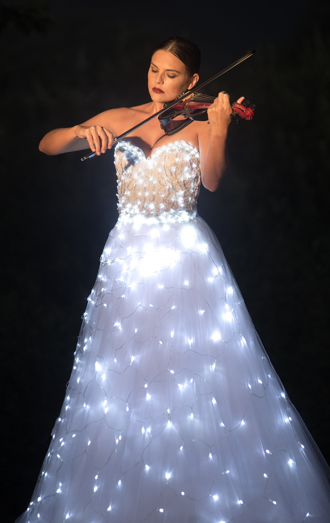 Woman playing violin wearing a white dress decorated with glowing string lights against a dark background.